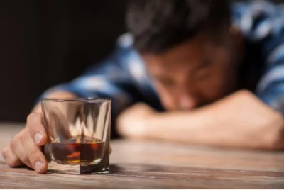 Man with his head on the table rests his hand on a glass of alcohol