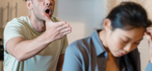 A man who has recently had alcohol appears to be angrily shouting and gesturing towards a woman, who looks distressed and is turning away with her head down.