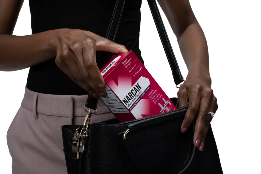 The image shows a person holding a Narcan nasal spray box, which is a medication used to reverse opioid overdoses, including those caused by potent substances like fentanyl. This visual emphasizes the importance of carrying naloxone as a life-saving measure in the ongoing overdose crisis.
